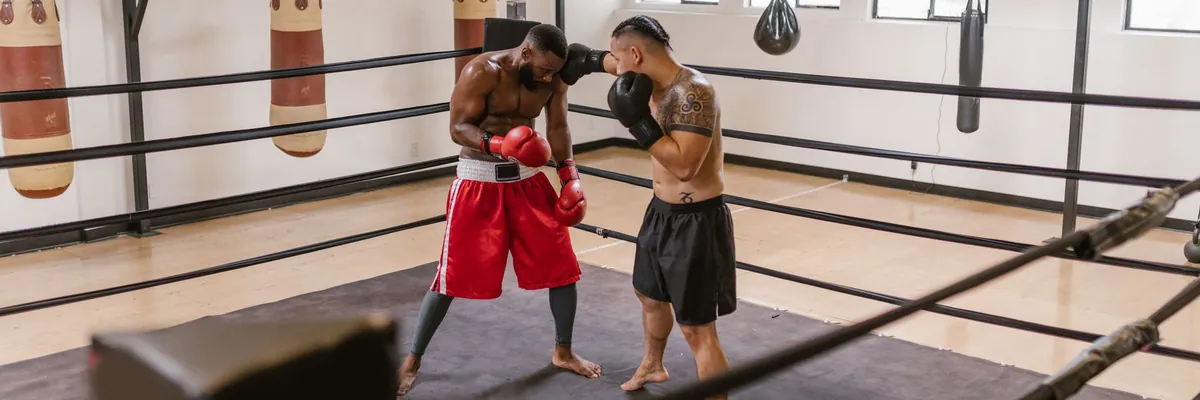 Two fighters sparring in a boxing ring with heavy bags behind